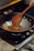 A wooden spoon is stirring a mixture of spices and food in a black wok on a gas stove. The food appears to be sautéing with visible bits of red and brown ingredients. The setting is a kitchen with a focus on cooking.