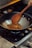 A wooden spoon is stirring a mixture of spices and food in a black wok on a gas stove. The food appears to be sautéing with visible bits of red and brown ingredients. The setting is a kitchen with a focus on cooking.
