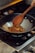 A wooden spoon is stirring a mixture of spices and food in a black wok on a gas stove. The food appears to be sautéing with visible bits of red and brown ingredients. The setting is a kitchen with a focus on cooking.