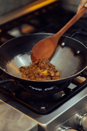A wooden spoon is stirring a mixture of spices and food in a black wok on a gas stove. The food appears to be sautéing with visible bits of red and brown ingredients. The setting is a kitchen with a focus on cooking.