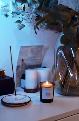 A lit candle in a glass jar sits on a white surface beside some cylindrical candles and a photograph in the background. There is an incense stick placed on a ceramic holder, and a large, clear vase containing leafy branches stands nearby.