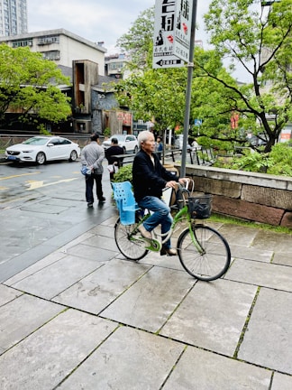 An elderly person rides a bicycle along a paved sidewalk beside a road with passing cars. In the background, trees and urban buildings are visible, along with a few pedestrians walking. A sign with Chinese characters is posted prominently on a pole.