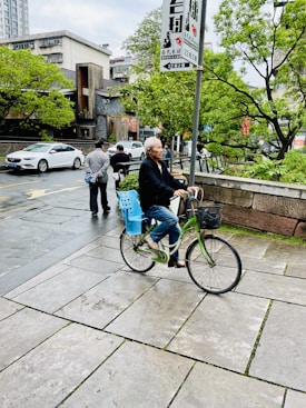 An elderly person rides a bicycle along a paved sidewalk beside a road with passing cars. In the background, trees and urban buildings are visible, along with a few pedestrians walking. A sign with Chinese characters is posted prominently on a pole.