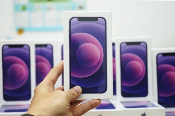 A hand is holding a boxed smartphone with a stylized purple and pink circle design on the front. Multiple identical smartphone boxes are displayed in the background.