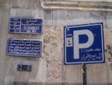 Street signs in Arabic and English are displayed on a wall with weathered surfaces. The largest sign shows a parking symbol 'P' with Arabic writing beneath it. Other signs provide information about the street, including historical references and street numbers.