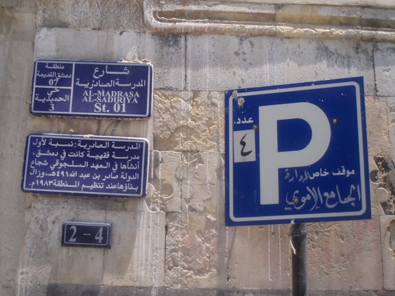 Street signs in Arabic and English are displayed on a wall with weathered surfaces. The largest sign shows a parking symbol 'P' with Arabic writing beneath it. Other signs provide information about the street, including historical references and street numbers.