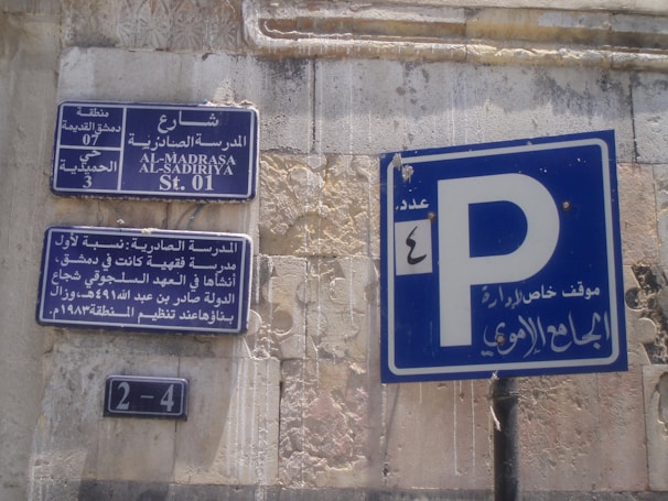 Street signs in Arabic and English are displayed on a wall with weathered surfaces. The largest sign shows a parking symbol 'P' with Arabic writing beneath it. Other signs provide information about the street, including historical references and street numbers.