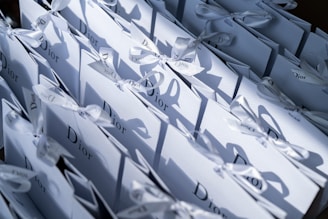 A collection of elegantly arranged white gift bags with the Dior logo, each tied with a neat bow. The bags are lined up closely together in an orderly fashion, casting soft shadows.