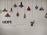 A wall displaying a series of decorative lamp illustrations hanging from black cords. The lamps vary in color and design, including shades of red, teal, brown, and black with floral patterns. Below the lamps, large text spells 'HOPE', accompanied by a poem about hope and resilience.