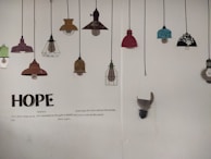 A wall displaying a series of decorative lamp illustrations hanging from black cords. The lamps vary in color and design, including shades of red, teal, brown, and black with floral patterns. Below the lamps, large text spells 'HOPE', accompanied by a poem about hope and resilience.