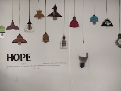 A wall displaying a series of decorative lamp illustrations hanging from black cords. The lamps vary in color and design, including shades of red, teal, brown, and black with floral patterns. Below the lamps, large text spells 'HOPE', accompanied by a poem about hope and resilience.