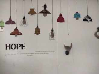 A wall displaying a series of decorative lamp illustrations hanging from black cords. The lamps vary in color and design, including shades of red, teal, brown, and black with floral patterns. Below the lamps, large text spells 'HOPE', accompanied by a poem about hope and resilience.