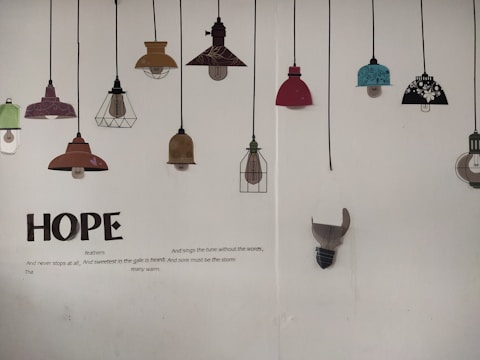 A wall displaying a series of decorative lamp illustrations hanging from black cords. The lamps vary in color and design, including shades of red, teal, brown, and black with floral patterns. Below the lamps, large text spells 'HOPE', accompanied by a poem about hope and resilience.