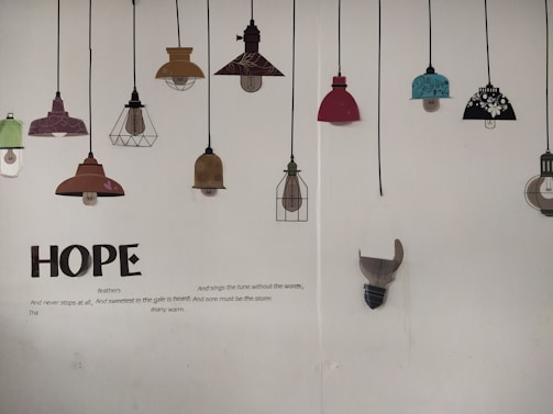 A wall displaying a series of decorative lamp illustrations hanging from black cords. The lamps vary in color and design, including shades of red, teal, brown, and black with floral patterns. Below the lamps, large text spells 'HOPE', accompanied by a poem about hope and resilience.
