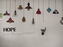 A wall displaying a series of decorative lamp illustrations hanging from black cords. The lamps vary in color and design, including shades of red, teal, brown, and black with floral patterns. Below the lamps, large text spells 'HOPE', accompanied by a poem about hope and resilience.