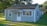 A small, single-story house with gray siding and a dark shingled roof. It features a white door with multiple panes of glass and has striped awnings over the front windows. The house is surrounded by a neatly trimmed lawn and several mature trees are visible in the background.
