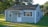 A small, single-story house with gray siding and a dark shingled roof. It features a white door with multiple panes of glass and has striped awnings over the front windows. The house is surrounded by a neatly trimmed lawn and several mature trees are visible in the background.
