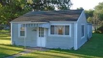 A small, single-story house with gray siding and a dark shingled roof. It features a white door with multiple panes of glass and has striped awnings over the front windows. The house is surrounded by a neatly trimmed lawn and several mature trees are visible in the background.