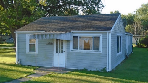 A small, single-story house with gray siding and a dark shingled roof. It features a white door with multiple panes of glass and has striped awnings over the front windows. The house is surrounded by a neatly trimmed lawn and several mature trees are visible in the background.