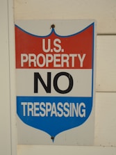 A sign with a red, white, and blue shield design displays the words 'U.S. Property No Trespassing' in bold letters. It is mounted on a light-colored wooden wall.