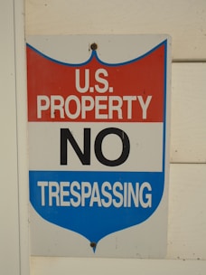 A sign with a red, white, and blue shield design displays the words 'U.S. Property No Trespassing' in bold letters. It is mounted on a light-colored wooden wall.