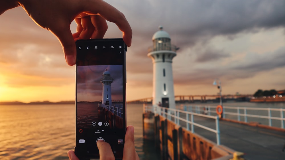 Person mit schwarzem Smartphone beim Fotografieren des weißen Leuchtturms während des Sonnenuntergangs