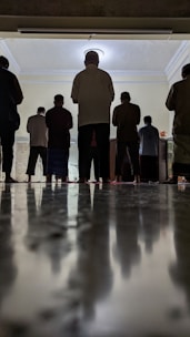 A group of people are standing in a room, viewed from a low angle that emphasizes the reflective flooring. The individuals appear engaged in an activity, possibly a prayer or meditation session, based on their posture. The room is softly lit, adding a serene atmosphere.