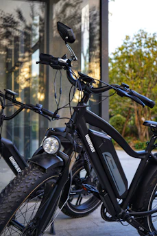 A close-up view of two black e-bikes parked near a glass window reflecting trees. Both bikes have thick tires and the brand name 'Himiway' visibly printed on the frames. The background shows foliage and a partly cloudy sky.