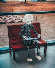 A humanoid robot with a white head and realistic design is seated on a wooden bench indoors. The robot appears to be engaging with a tablet device. Behind the bench, a large window provides a view of a grassy outdoor area with what looks like an art installation or seating arrangement.