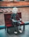A humanoid robot with a white head and realistic design is seated on a wooden bench indoors. The robot appears to be engaging with a tablet device. Behind the bench, a large window provides a view of a grassy outdoor area with what looks like an art installation or seating arrangement.