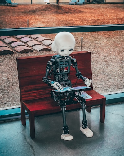 A humanoid robot with a white head and realistic design is seated on a wooden bench indoors. The robot appears to be engaging with a tablet device. Behind the bench, a large window provides a view of a grassy outdoor area with what looks like an art installation or seating arrangement.