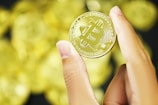 A hand holding a gold-colored coin with a Bitcoin symbol embossed on its surface. The background is filled with blurred similar coins, creating a shiny and luxurious appearance.