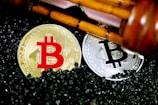 Golden and silver Bitcoin coins are placed on a surface covered with small black granules. Above the coins, a wooden gavel is positioned diagonally, suggesting a theme of cryptocurrency regulation or legal matters.