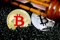 Golden and silver Bitcoin coins are placed on a surface covered with small black granules. Above the coins, a wooden gavel is positioned diagonally, suggesting a theme of cryptocurrency regulation or legal matters.