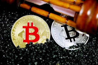 Golden and silver Bitcoin coins are placed on a surface covered with small black granules. Above the coins, a wooden gavel is positioned diagonally, suggesting a theme of cryptocurrency regulation or legal matters.