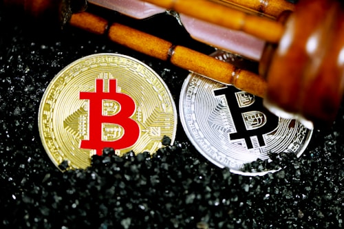 Golden and silver Bitcoin coins are placed on a surface covered with small black granules. Above the coins, a wooden gavel is positioned diagonally, suggesting a theme of cryptocurrency regulation or legal matters.