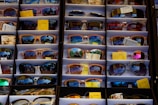 A display of various sunglasses neatly arranged in rows. Each pair reflects a vibrant outdoor scene, likely with trees and a bright sky. The sunglasses vary in shape, size, and lens color, and some have price tags attached.