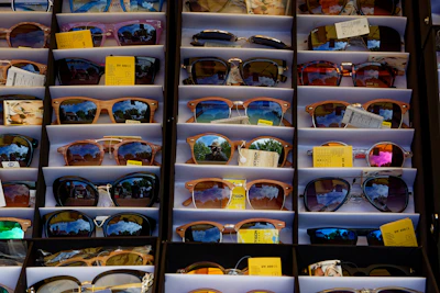 A vibrant display of sunglasses in various colors and shapes on a wooden table with sunlight filtering through leaves.