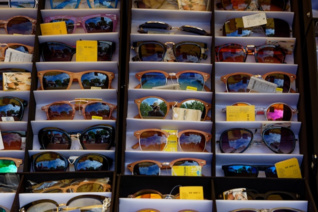 A display of various sunglasses neatly arranged in rows. Each pair reflects a vibrant outdoor scene, likely with trees and a bright sky. The sunglasses vary in shape, size, and lens color, and some have price tags attached.