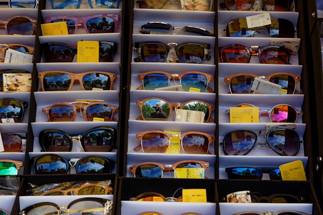 A display of various sunglasses neatly arranged in rows. Each pair reflects a vibrant outdoor scene, likely with trees and a bright sky. The sunglasses vary in shape, size, and lens color, and some have price tags attached.