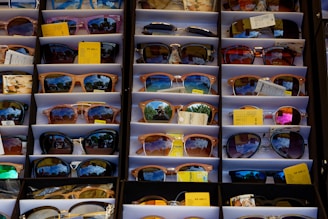 A display of various sunglasses neatly arranged in rows. Each pair reflects a vibrant outdoor scene, likely with trees and a bright sky. The sunglasses vary in shape, size, and lens color, and some have price tags attached.
