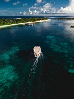 Aerial view of a boat moving through clear, turquoise waters with a lush island in the distance. The sky is partly cloudy with hints of blue and the water shows varying shades of blue and green, indicating changes in depth. Over-water bungalows are visible on the right side of the island.