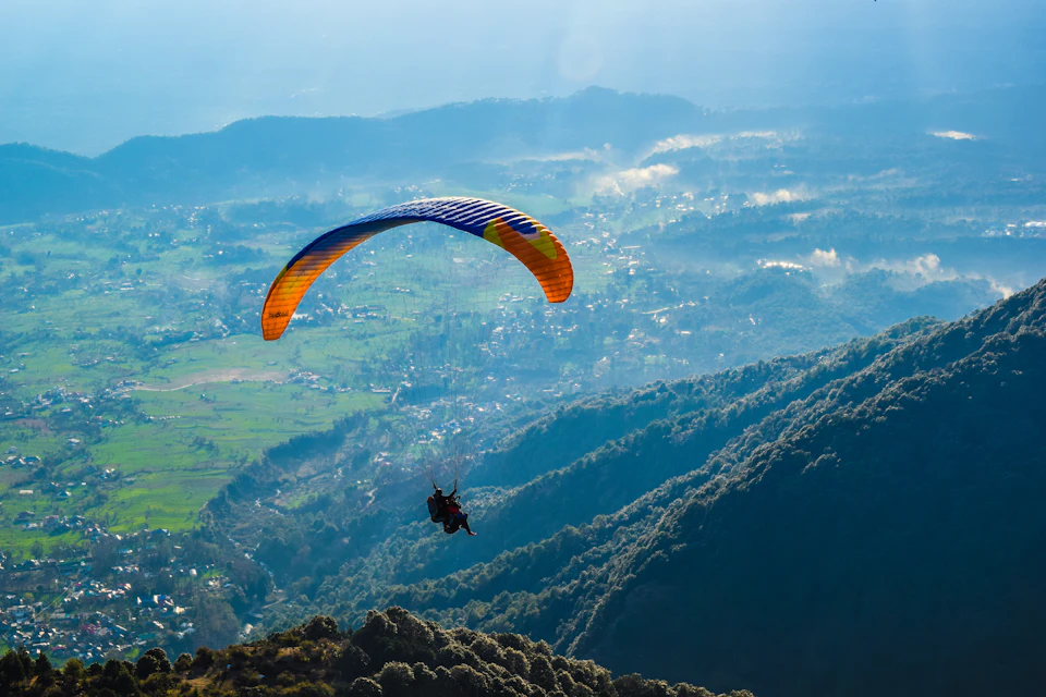 The image depicts a paraglider suspended in mid-air, soaring over a verdant valley below. The paraglider is painted in vibrant hues of orange and blue, with a yellow parachute billowing behind it. Below the paraglider's feet, a lush green valley stretches out, dotted with trees that add depth to the scene. In the distance, mountains rise against the horizon, their peaks shrouded in mist. The sky above is a clear blue canvas, providing a stunning backdrop for this airborne adventure.