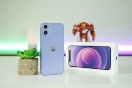 A purple smartphone is standing upright on a white surface next to a small potted plant. Behind it, there is a white box displaying an image of the smartphone screen. In the background, a blurred figurine and colorful lighting create a soft ambiance.
