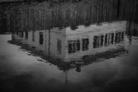 A reflective image of a building is seen on the surface of rippling water. The reflection creates a mirror-like effect, capturing windows and architectural details against a textured, wavy background.