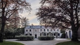 A large, elegant mansion surrounded by trees with lush foliage, featuring a grand driveway leading to the entrance. The building has several windows, a stone facade, and is framed by well-kept greenery.