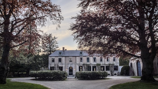 A large, elegant mansion surrounded by trees with lush foliage, featuring a grand driveway leading to the entrance. The building has several windows, a stone facade, and is framed by well-kept greenery.