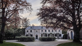 A large, elegant mansion surrounded by trees with lush foliage, featuring a grand driveway leading to the entrance. The building has several windows, a stone facade, and is framed by well-kept greenery.