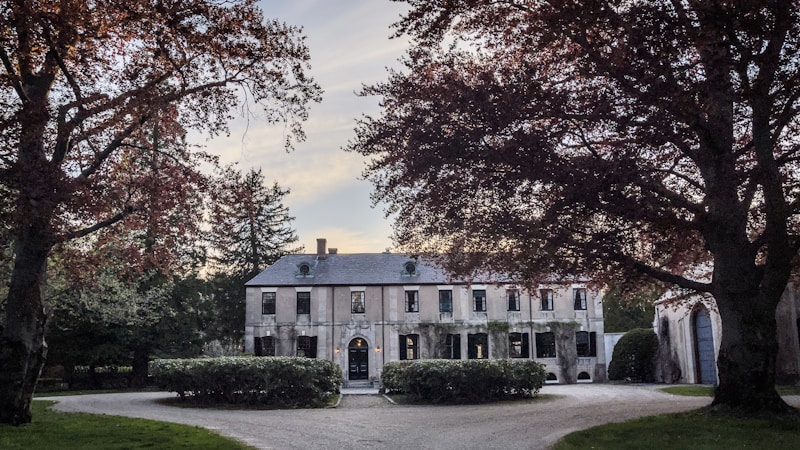 A large, elegant mansion surrounded by trees with lush foliage, featuring a grand driveway leading to the entrance. The building has several windows, a stone facade, and is framed by well-kept greenery.