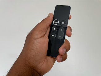 A hand holds a sleek, black remote control featuring several buttons including menu, play/pause, and volume controls. The remote has a modern design with a glossy finish.